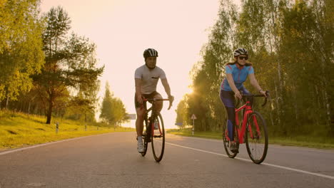 Un-Hombre-Y-Una-Mujer-Montan-Bicicletas-Deportivas-En-La-Carretera-Al-Atardecer-Con-Equipo-Y-Cascos-Protectores-En-Cámara-Lenta-A-120-Fps.